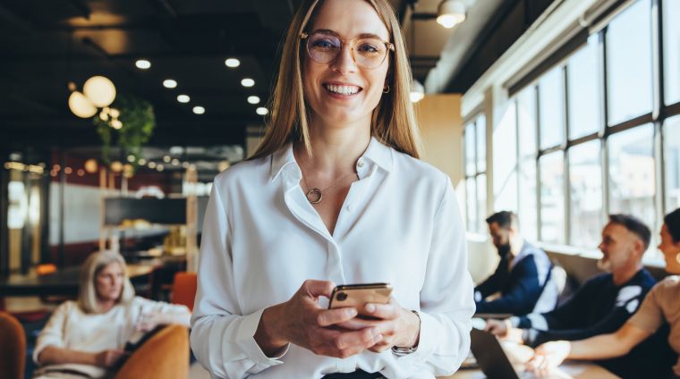 Young businesswoman holding a smartphone in a co-working space. Happy young businesswoman smiling at the camera while standing in a modern workplace. Female entrepreneur sending a text message.